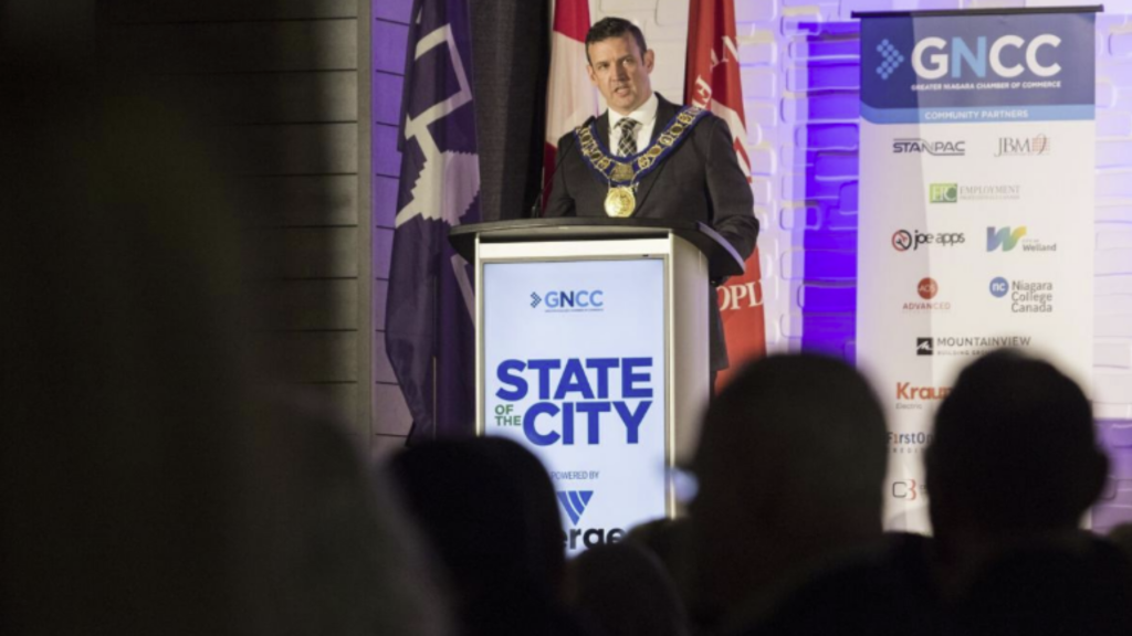 A person wearing a ceremonial chain speaks at a podium labeled "State of the City." They stand beside flags and a banner with sponsor logos. The setting suggests a formal event.