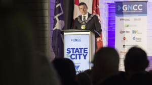 A person wearing a ceremonial chain speaks at a podium labeled "State of the City." They stand beside flags and a banner with sponsor logos. The setting suggests a formal event.