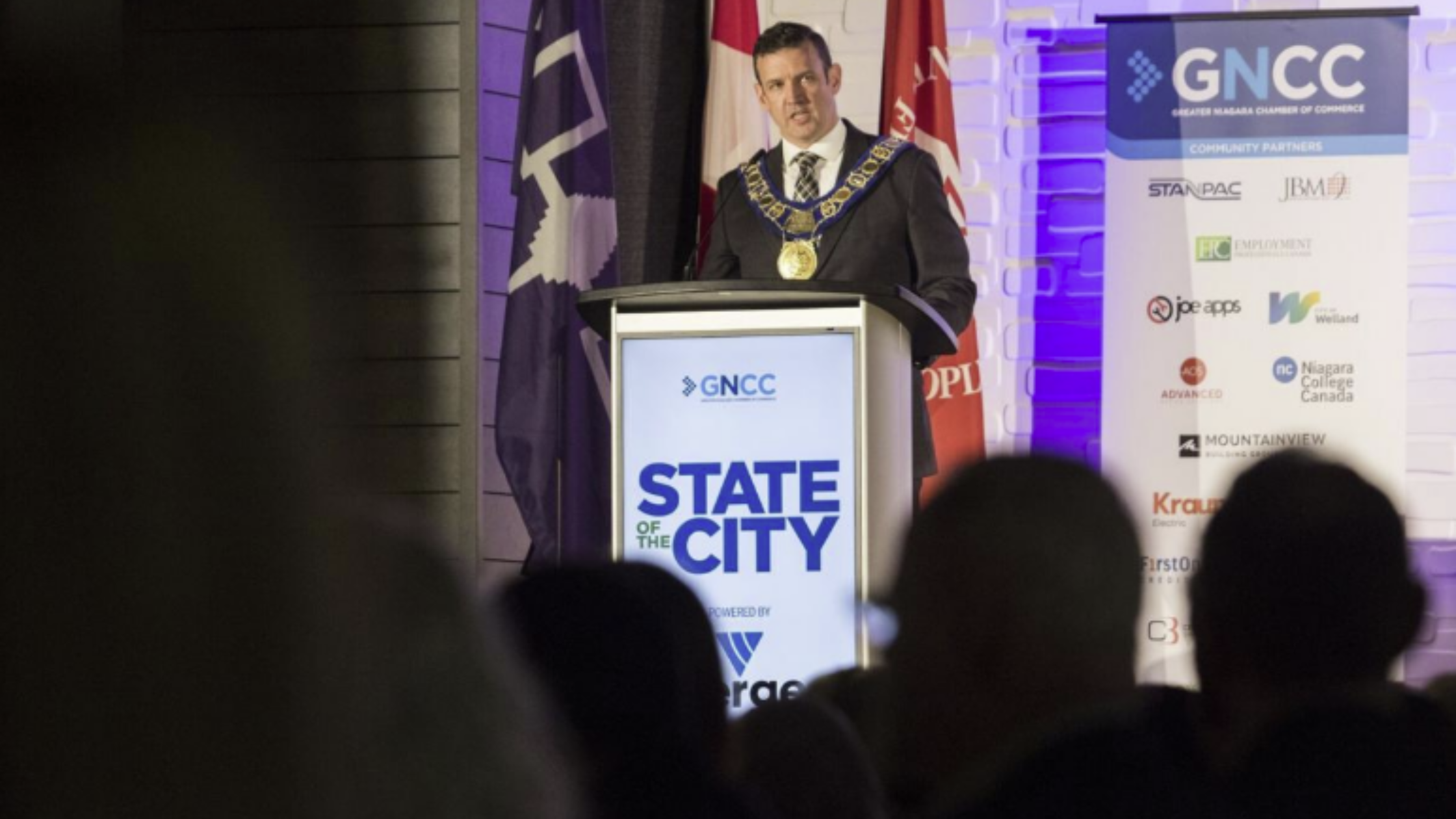 A person wearing a ceremonial chain speaks at a podium labeled "State of the City." They stand beside flags and a banner with sponsor logos. The setting suggests a formal event.