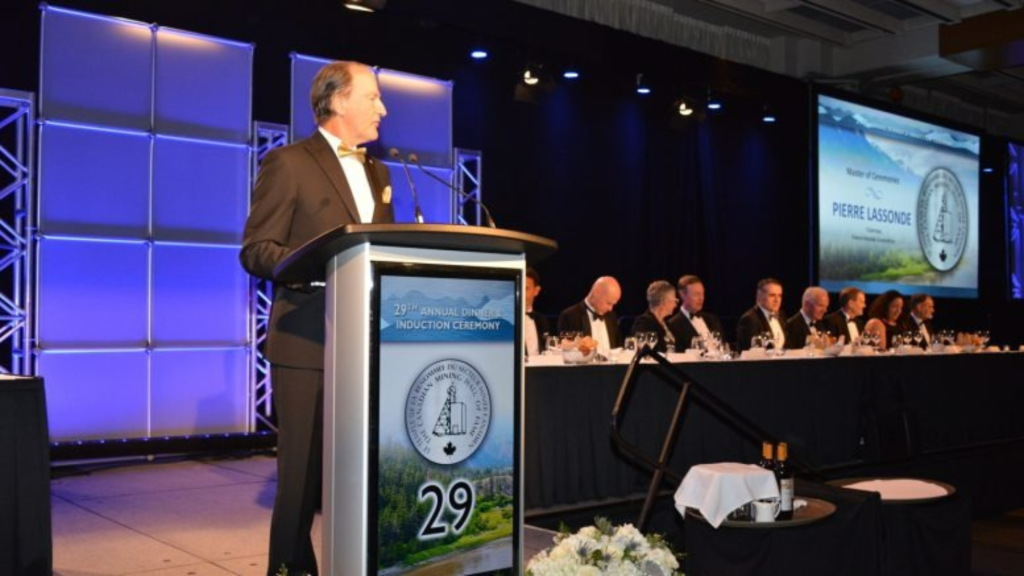 Speaker delivering a presentation at a formal business awards and induction ceremony with attendees seated on stage during a banquet event.