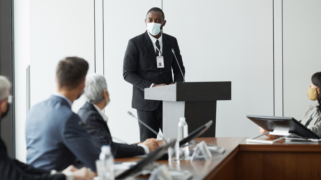 Business professional wearing a face mask delivers a presentation at a podium during a corporate meeting while attendees listen from a conference table.