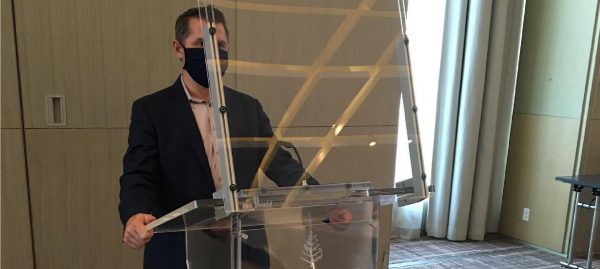 Person with COVID-19 face mask standing behind a podium with an acrylic barrier